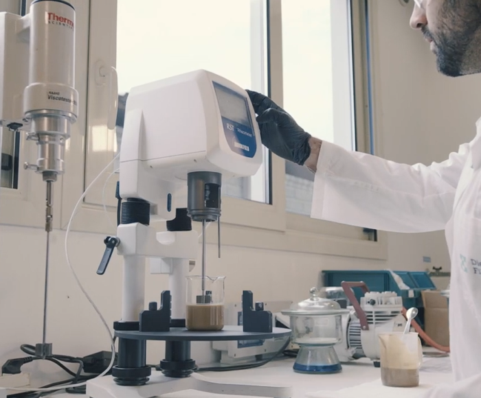 técnico del laboratorio Diemme® Filtration realizando un ensayo de sedimentación en probeta junto a una celda de filtración piloto, con muestra de lodo real del cliente y registro de datos en pantalla