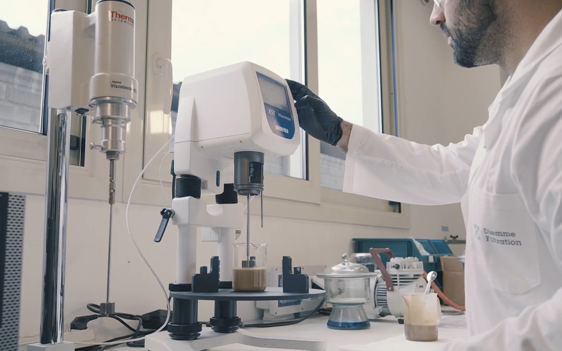 técnico del laboratorio Diemme® Filtration realizando un ensayo de sedimentación en probeta junto a una celda de filtración piloto, con muestra de lodo real del cliente y registro de datos en pantalla